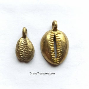 May include: Two gold-colored pendants. One is a small, textured teardrop shape. The other is a larger, oval pendant with a slit down the center, resembling a shell. Both have small loops at the top for hanging. The website address "GhanaTreasures.com" is visible.