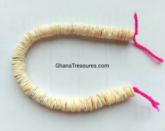 May include: A strand of white shell beads strung on pink thread. The beads are flat and round, and they are arranged in a circle. The text "GhanaTreasures.com" is visible in the image.