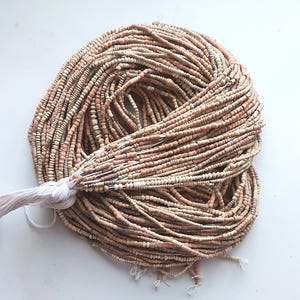 Peut inclure: Une grande collection de nombreux brins de petites perles cylindriques aux tons terreux, dans des nuances de beige, de brun clair et de sable. Les brins sont rassemblés et noués avec une ficelle blanche à une extrémité, formant un cercle.