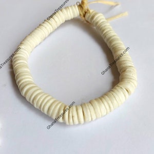 以下が含まれることがあります： 小さな丸い平らなビーズで作られた白いブレスレット。ビーズは天然繊維の紐で繋げられています。