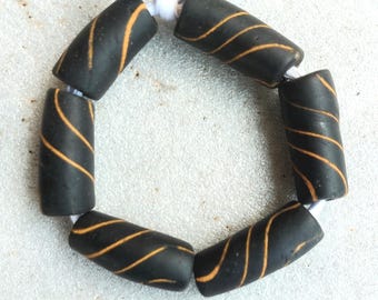 May include: A strand of black barrel beads with orange swirl designs. The beads are strung together with white string, forming a circular shape. The beads are approximately 1 inch long.