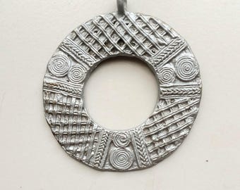 May include: A silver-colored pendant with a circular design and a central hole. The pendant features intricate patterns, including spirals, woven textures, and diagonal lines. It has a small loop at the top for hanging.