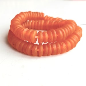 以下が含まれることがあります： オレンジ色のガラスビーズのネックレス。ビーズはそれぞれ、中央に穴が開いた小さな平らな円盤形をしています。ビーズは糸に通して長いネックレスになっています。