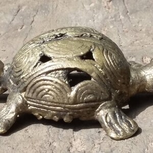 May include: A small, gold-colored metal turtle figurine with intricate details on its shell. The turtle is lying on its belly with its head turned to the side.