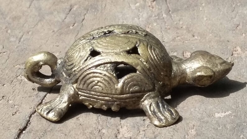 May include: A small, gold-colored metal turtle figurine with intricate details on its shell. The turtle is lying on its belly with its head turned to the side.
