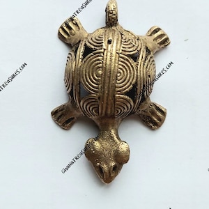May include: A gold-colored metal turtle pendant with a detailed, circular pattern on its shell. The pendant has a loop at the top for hanging.