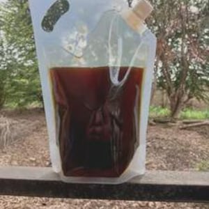 Può includere: Una busta flessibile e trasparente in plastica, capace di stare in piedi, riempita con un liquido marrone scuro. La busta presenta un'apertura integrata per la mano in alto a sinistra e un tappo a vite bianco nell'angolo in alto a destra.