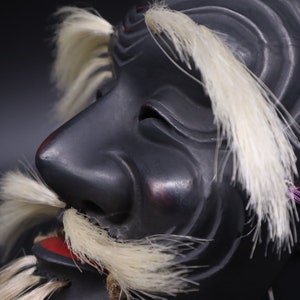 May include: A black wooden mask with white feathers and a red mouth. The mask has a long nose and a furrowed brow, giving it a fierce expression.
