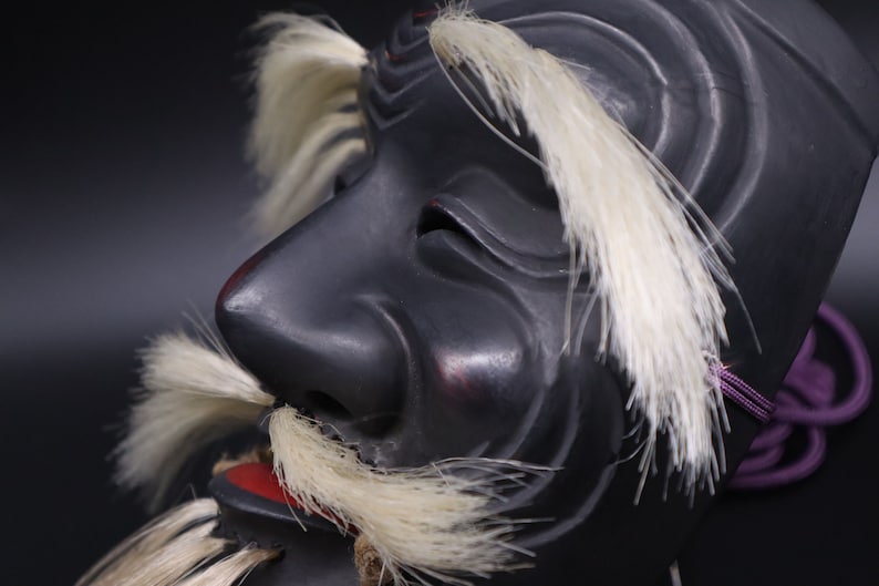 May include: A black wooden mask with white feathers and a red mouth. The mask has a long nose and a furrowed brow, giving it a fierce expression.