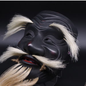 May include: A black wooden mask with a white beard and mustache. The mask has a carved face with a serious expression and red painted lips.