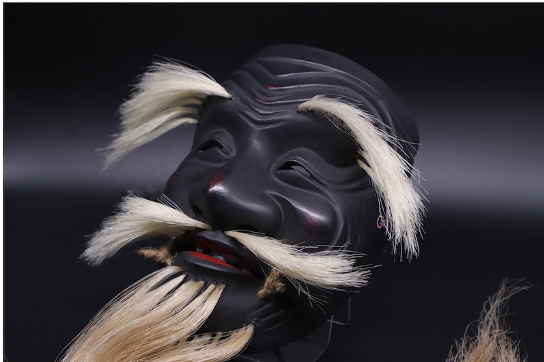 May include: A black wooden mask with a white beard and mustache. The mask has a carved face with a serious expression and red painted lips.