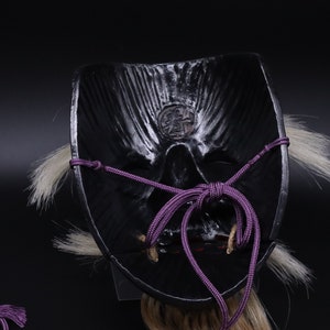 May include: Black Japanese Oni mask with a purple cord tied around the back. The mask has a detailed face with a carved mouth and eyes. The mask is decorated with white hair.
