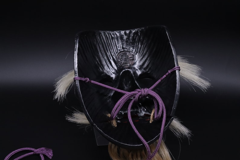 May include: Black Japanese Oni mask with a purple cord tied around the back. The mask has a detailed face with a carved mouth and eyes. The mask is decorated with white hair.