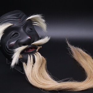 May include: A black wooden mask with a long white beard and mustache. The mask has a carved face with closed eyes and a slightly open mouth. The beard and mustache are made of real hair.
