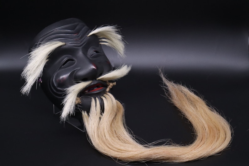 May include: A black wooden mask with a long white beard and mustache. The mask has a carved face with closed eyes and a slightly open mouth. The beard and mustache are made of real hair.