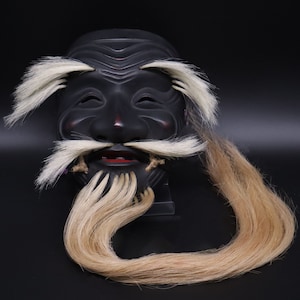 May include: A black wooden mask with a long, flowing blonde beard. The mask has a carved face with a wide mouth and closed eyes. The beard is made of real hair and is attached to the mask.