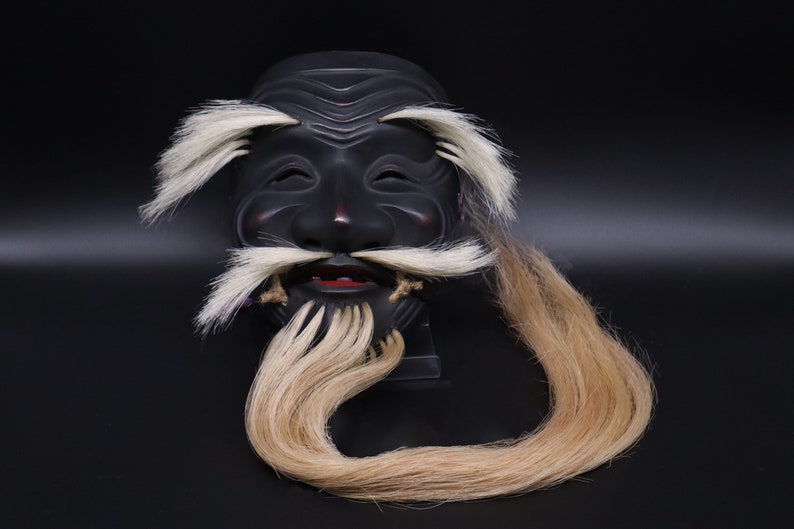 May include: A black wooden mask with a long, flowing blonde beard. The mask has a carved face with a wide mouth and closed eyes. The beard is made of real hair and is attached to the mask.