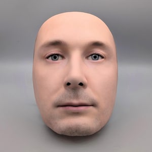 Puede incluir: Una máscara realista de tamaño natural de la cara de un hombre. La máscara tiene un tono de piel pálido, ojos azules y cabeza calva. La cara presenta un bigote corto y barba incipiente. El fondo es de color gris claro sólido.