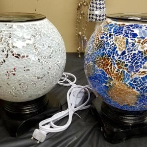 May include: Two round mosaic glass essential oil diffusers with black bases and electrical cords. One diffuser is white with silver accents and the other is blue with gold and brown accents.
