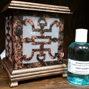 May include: A black and copper metal oil diffuser with a decorative design and a clear glass bottle of scented oil labeled "Scented Lizard Poppy Flowers Liquid burning oils".