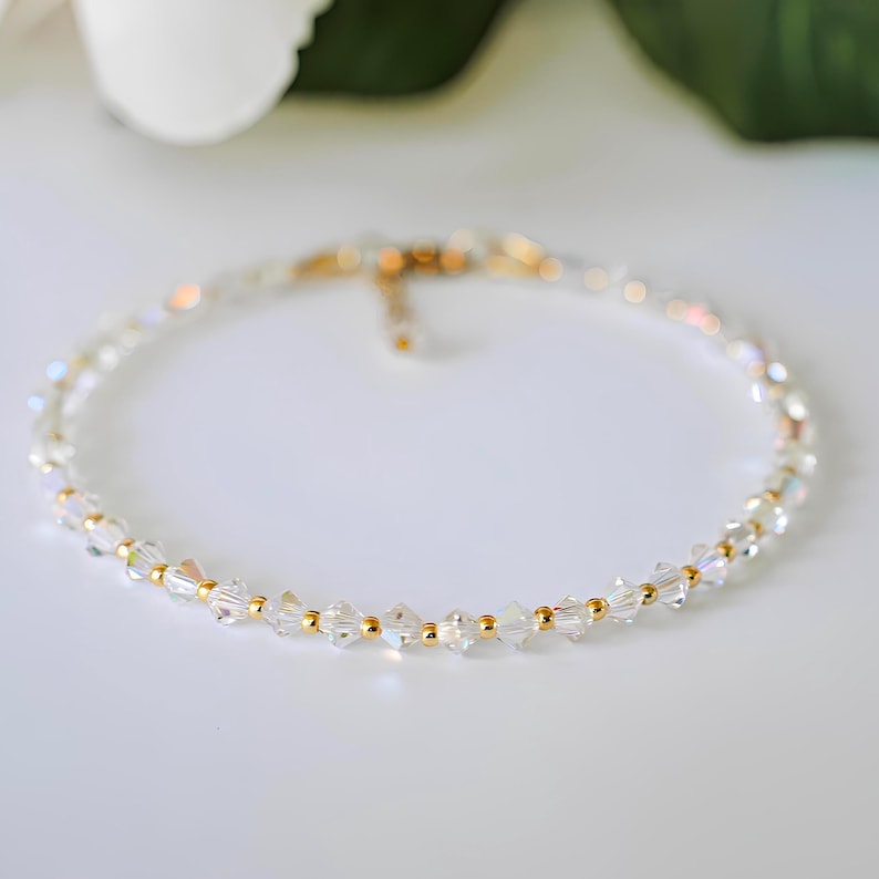May include: A delicate, clear crystal anklet with gold-colored beads. The anklet is made of small, faceted crystals strung together with gold-colored beads. The anklet is displayed on a white surface, with a blurred background of white and green.