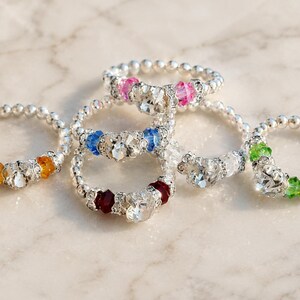 May include: A collection of beaded rings with silver-colored bands and colorful gemstone accents. The rings feature clear, pink, blue, red, orange, and green gemstones. The rings are displayed on a light-colored, marble-like surface.