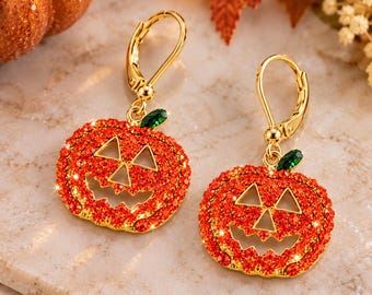 May include: A pair of gold-tone leverback earrings featuring pumpkin-shaped charms. Each pumpkin is encrusted with bright orange crystals, has cut-out eyes and a mouth, and is topped with a small, singular green crystal leaf.