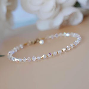 May include: Delicate bracelet featuring clear, faceted crystal beads strung on a gold-toned chain. The bracelet has small gold beads interspersed between the crystals. A small, round charm hangs near the clasp. The background is a soft, neutral color.