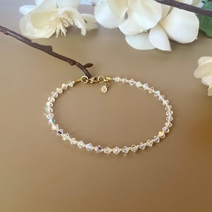 May include: A delicate bracelet featuring iridescent crystal beads strung on a gold-colored chain. The bracelet has a small charm near the clasp and is displayed on a neutral-toned surface, with white flowers in the background.