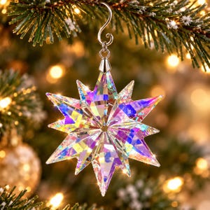 May include: A multi-pointed star ornament crafted from clear, faceted crystal with an iridescent finish that reflects rainbow colors. The star features a silver-toned metal cap and a curved hook for hanging, displaying sharp, geometric edges.