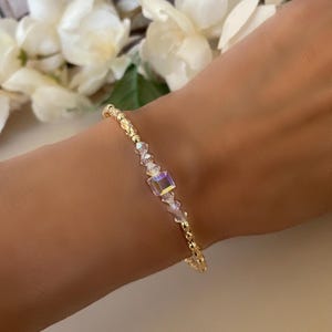 May include: A delicate gold bracelet with a central iridescent square crystal and smaller clear crystals. The bracelet features small gold beads and is worn on a wrist, showcasing its elegant design.