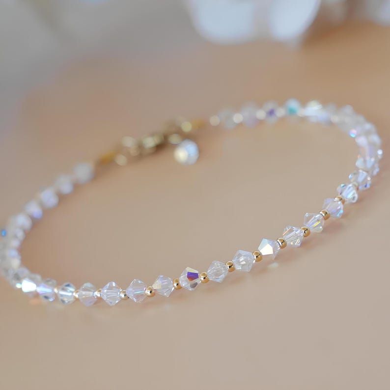 May include: Delicate bracelet featuring iridescent, faceted crystal beads interspersed with small gold-colored beads. The bracelet has a gold-colored clasp and a small charm. The crystals reflect a rainbow of colors, creating a subtle sparkle.