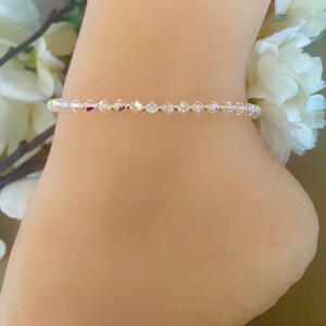 May include: A delicate ankle bracelet featuring small, clear, faceted beads strung on a gold-colored chain. The beads catch the light, creating a sparkling effect. The bracelet is shown on an ankle, suggesting it's a piece of jewelry.