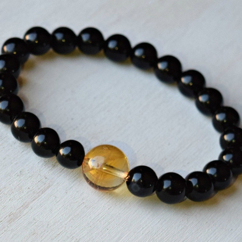 May include: A black beaded bracelet with a single amber bead. The bracelet is made of round, black beads and has a single, round, amber bead in the center.