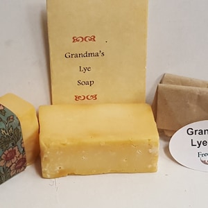 May include: Two bars of yellow lye soap, one wrapped in floral fabric and the other unwrapped. A small brown paper bag with a white oval sticker that reads "Grandma's Lye Soap Freed Family Farm" is in the background. A handwritten note reads "Grandma's Lye Soap".