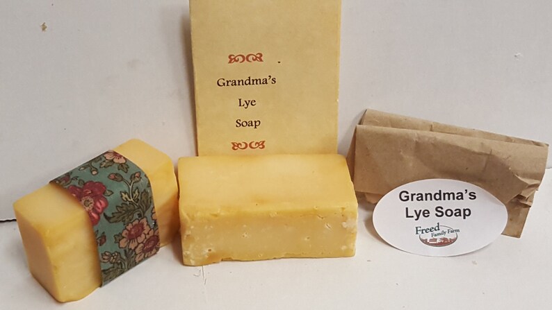 May include: Two bars of yellow lye soap, one wrapped in floral fabric and the other unwrapped. A small brown paper bag with a white oval sticker that reads "Grandma's Lye Soap Freed Family Farm" is in the background. A handwritten note reads "Grandma's Lye Soap".