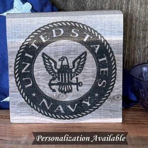 May include: A wooden block with the United States Navy emblem, featuring an eagle, anchor, and the words "United States Navy." The design is in black on a gray, weathered wood background. Text at the bottom reads "Personalization Available."