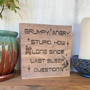 May include: A wooden sign with engraved text that reads "GRUMPY, ANGRY, STUPID, HOW LONG SINCE LAST SLEEP, QUESTION?" The sign has star and astronaut graphics. The sign is on a wooden surface.