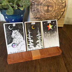 May include: Three tarot cards displayed in a wooden stand. The cards are titled The High Priestess, The Moon, and The Star. The High Priestess features a white tiger, The Moon shows pine trees, and The Star depicts a colorful star.