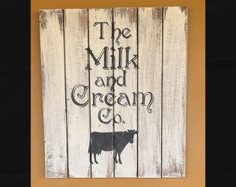 Peut inclure: Panneau en bois vieilli avec l'inscription "The Milk and Cream Co." en lettres noires. Une silhouette de vache est représentée en dessous. Le panneau a une finition blanchie à la chaux, lui donnant un aspect vintage.