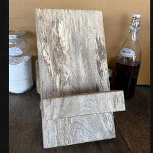 May include: A rustic wooden cookbook stand with a light gray and brown finish. The stand has a rectangular base and a back support. A glass bottle with a Vanilla label and a jar are in the background.