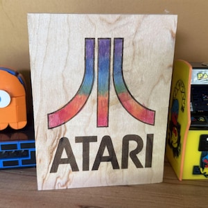 May include: A rectangular wooden plaque featuring the Atari logo with a rainbow-colored gradient design and the word ATARI in black lettering. To the left is an orange ghost figure and to the right is a miniature Pac-Man arcade cabinet.