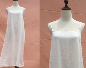Puede incluir: Un vestido lencero blanco de lino con finas tiras de espagueti. El vestido se exhibe en un maniquí con un soporte de madera. El vestido tiene un diseño sencillo y fluido, perfecto para el clima cálido o para combinar.
