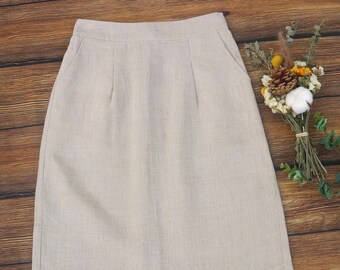 May include: A beige linen skirt with a classic A-line silhouette. The skirt features two front pockets and a tailored waistband. The fabric has a subtle texture, and the skirt is displayed on a wooden surface. A small bouquet of dried flowers is in the background.