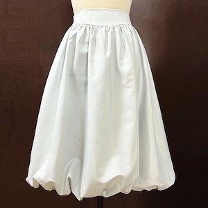 May include: A light gray, full-circle skirt with a gathered waistband. The skirt is made of a smooth, lightweight fabric and has a bubble hem.