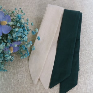 May include: Two fabric ribbon ties, one in a light beige color and the other in a dark green color. The ribbons are folded and arranged on a textured beige surface.