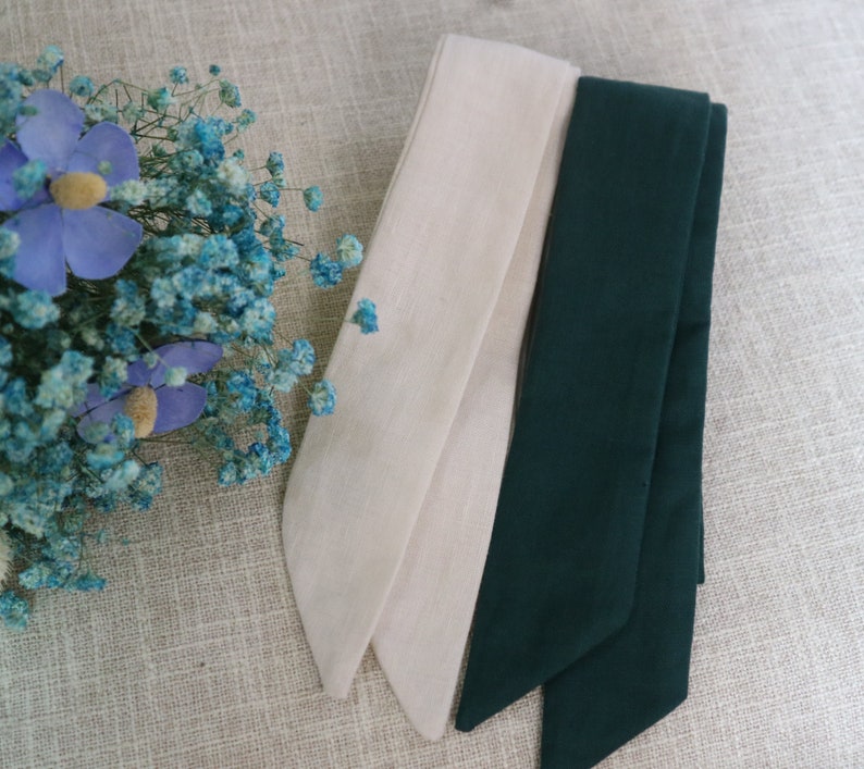 May include: Two fabric ribbon ties, one in a light beige color and the other in a dark green color. The ribbons are folded and arranged on a textured beige surface.