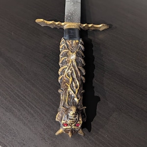 Puede incluir: Una espada decorativa con un mango dorado y una hoja plateada. El mango está tallado intrincadamente con una cara demoníaca y ojos rojos. La espada está sobre una superficie de madera oscura.