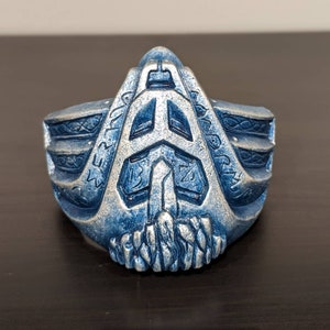 May include: A blue and silver face mask with a geometric design and runes. The mask has a pointed top and a jagged bottom.