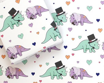 May include: White wrapping paper with a repeating pattern of green and purple dinosaurs wearing top hats and veils. The dinosaurs are holding bouquets of flowers. The background is filled with small hearts in various colours.